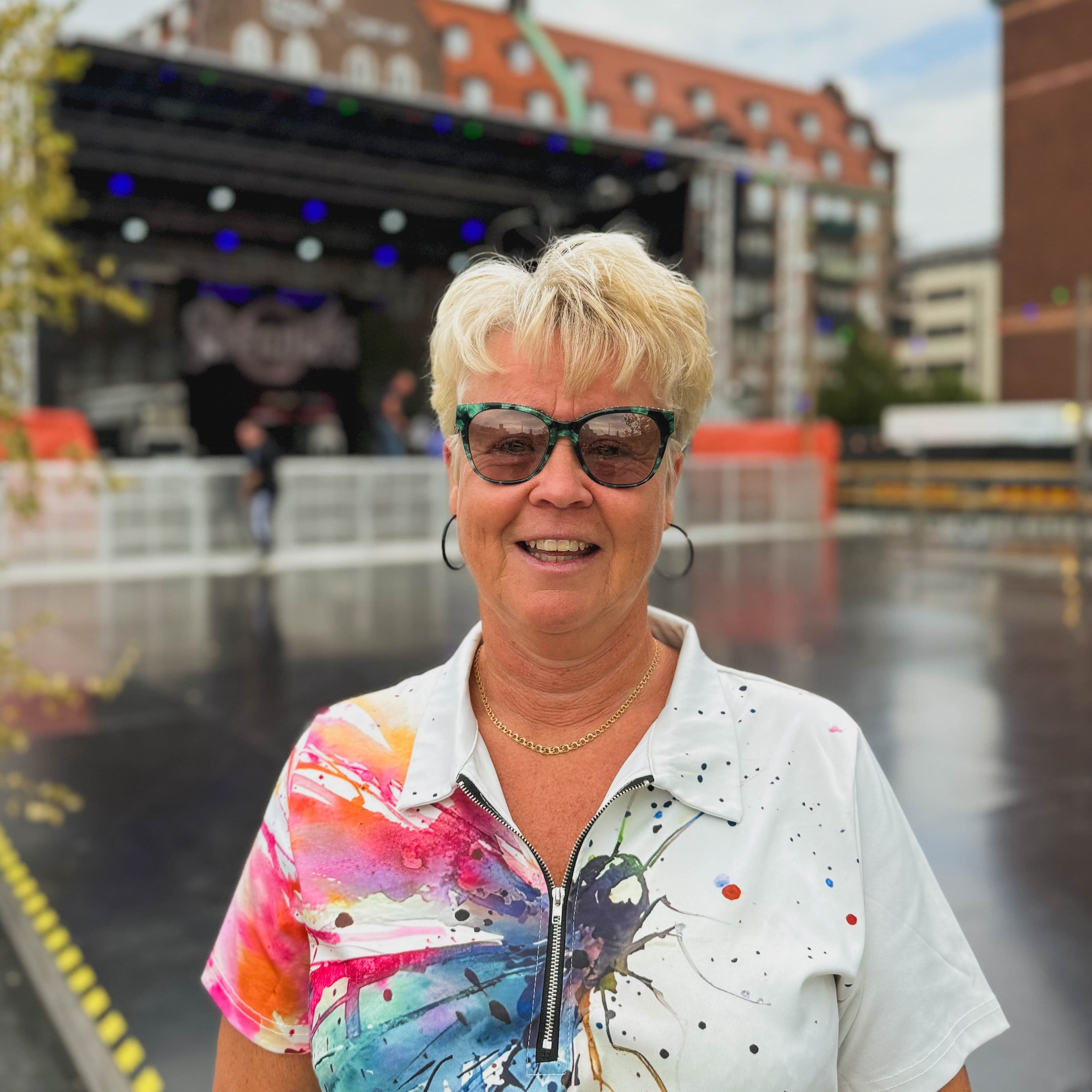 En kvinna med kort blont hår och stora solglasögon ler mot kameran. Hon bär en vit skjorta med färgstarka färgstänk i rosa, blått och lila. I bakgrunden syns en scenen Dansbanan.