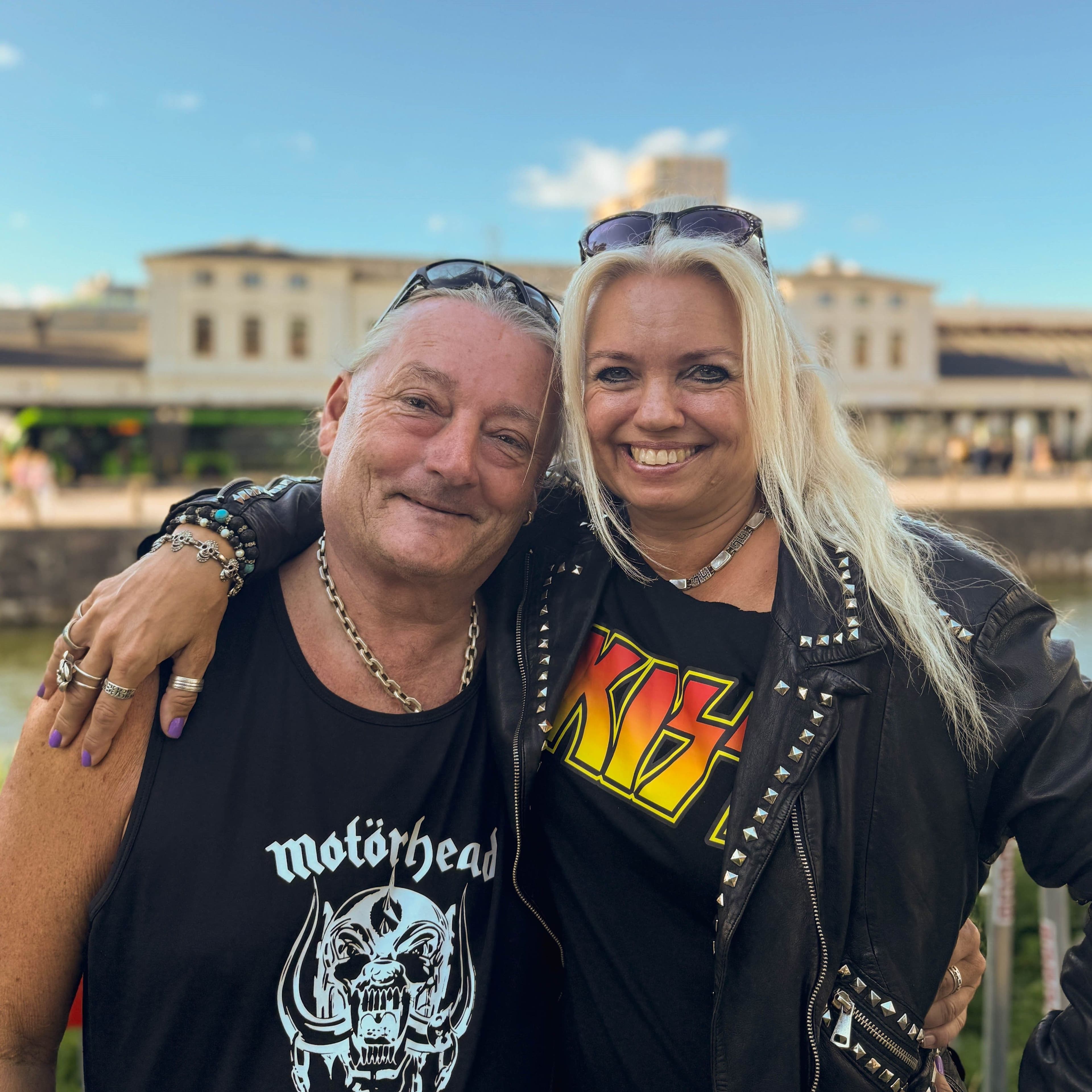 Ett leende par står utomhus med armen om varandra. Mannen bär ett svart linne med Motörhead-loggan och en silverkedja, medan kvinnan bär en svart läderjacka med nitar och en svart T-shirt med KISS-loggan. Bakom dem syns Malmö Centralstation och kanalen.