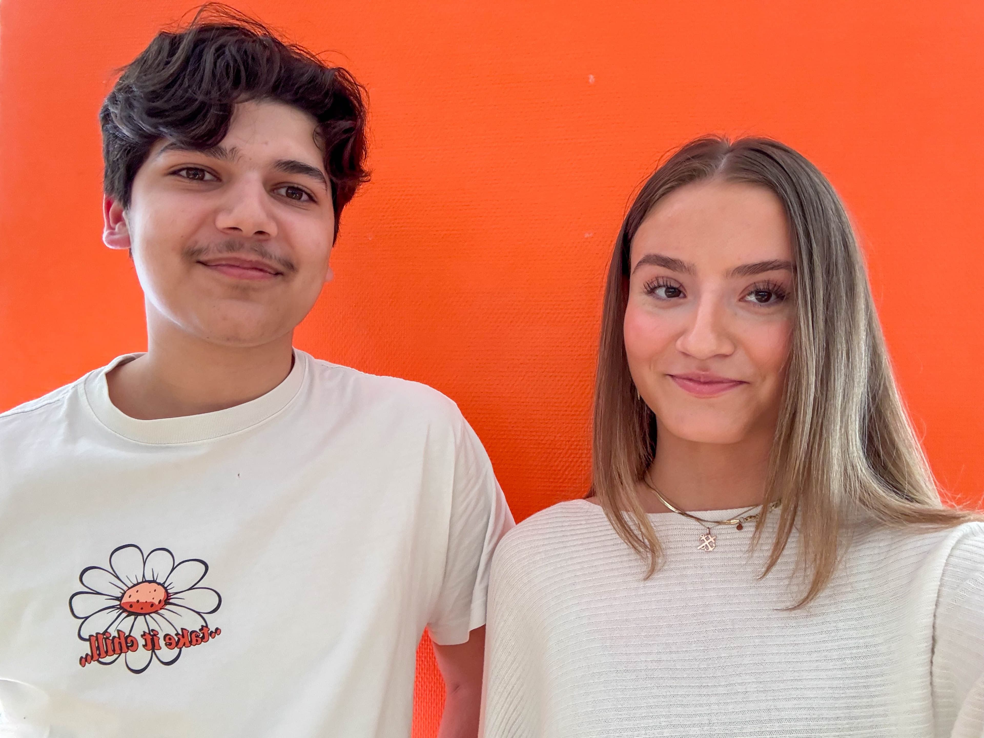 Två ungdomar står framför en orange vägg.